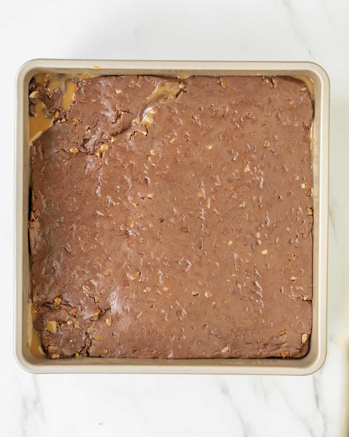 The unused brownie dough has been placed on top of the chocolate chip caramel layer in the square baking pan.