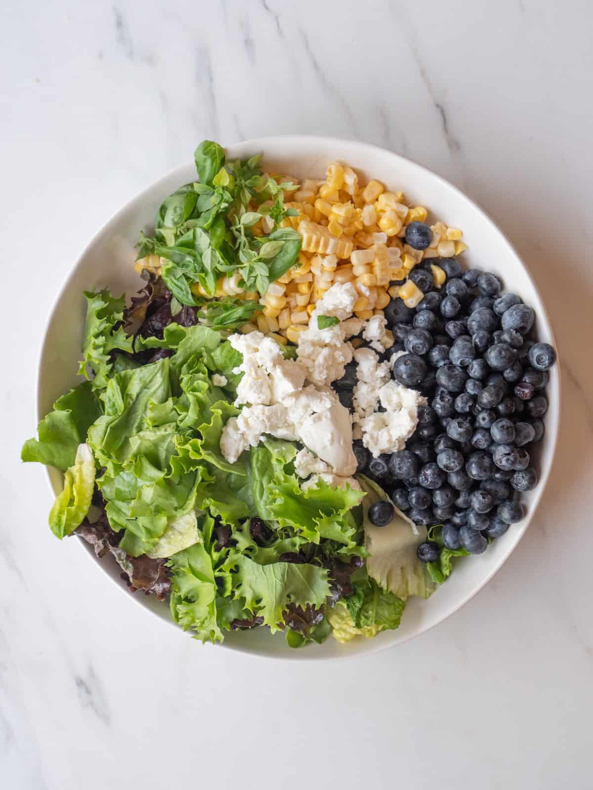 A large bowl with blueberries, goat cheese, basil leaves, lettuce and corn.