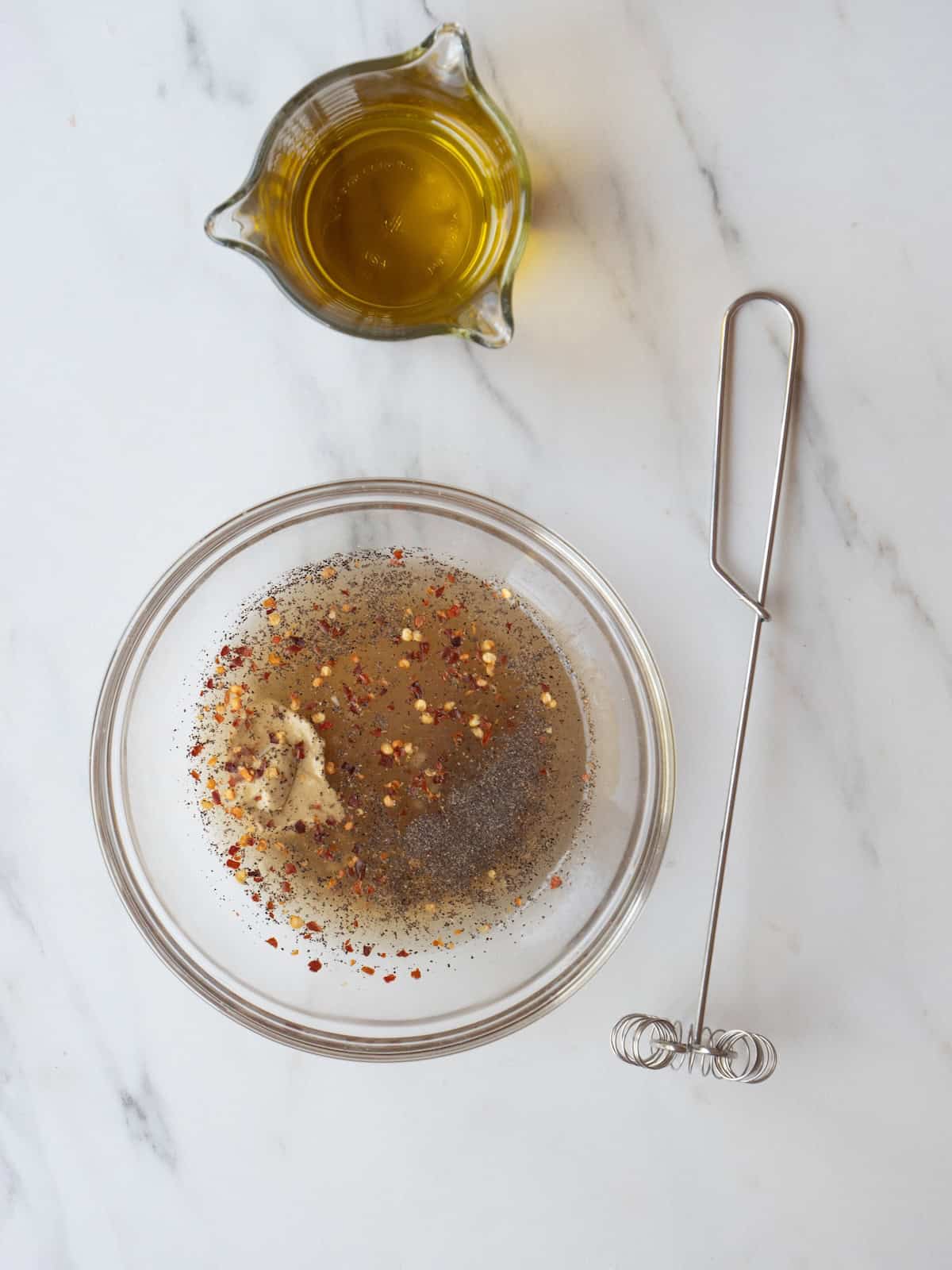 A small glass mixing bowl with all the ingredients for champagne vinaigrette along with a small whisk and a small jar of extra virgin olive oil.