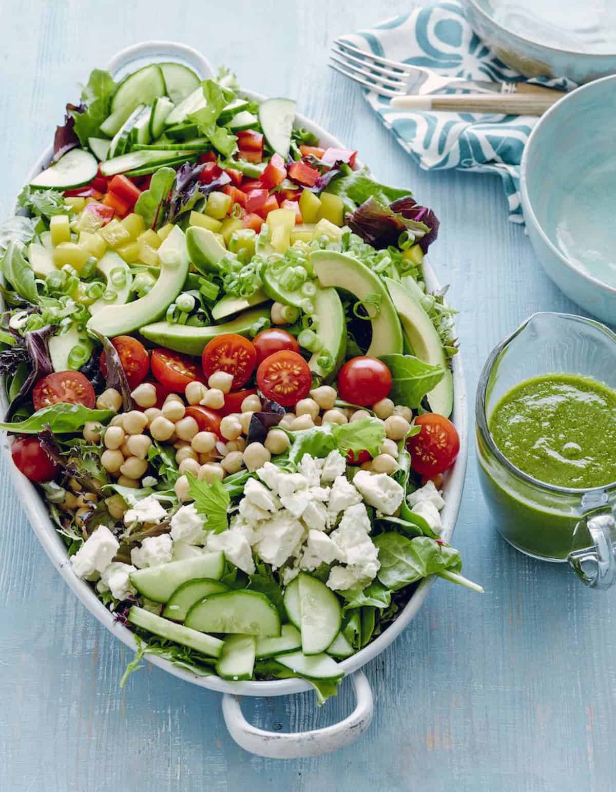 A bowl with loaded power salad containing a lot of veggies, chickpeas and cheese arranged in parallel rows on a bed of greens with basil vinaigrette on the side.