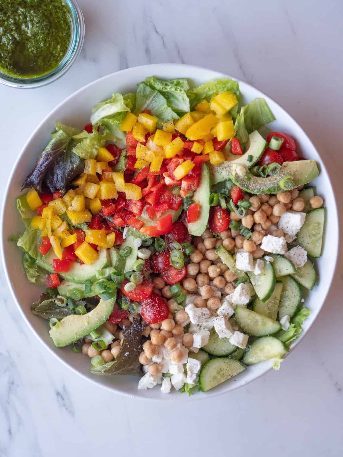 A bowl with loaded power salad containing a lot of veggies, chickpeas and cheese arranged in parallel rows on a bed of greens with basil vinaigrette on the side.