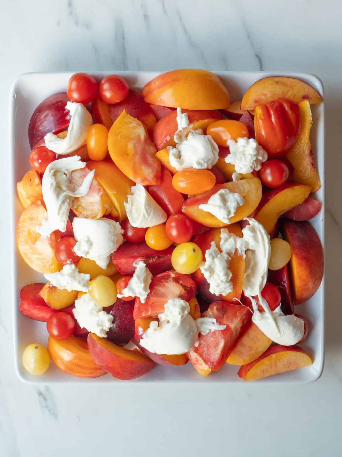 A plate with nectarines, plums, peaches and heirloom tomatoes topped with ripped burrata and salt and pepper, along with mint leaves.