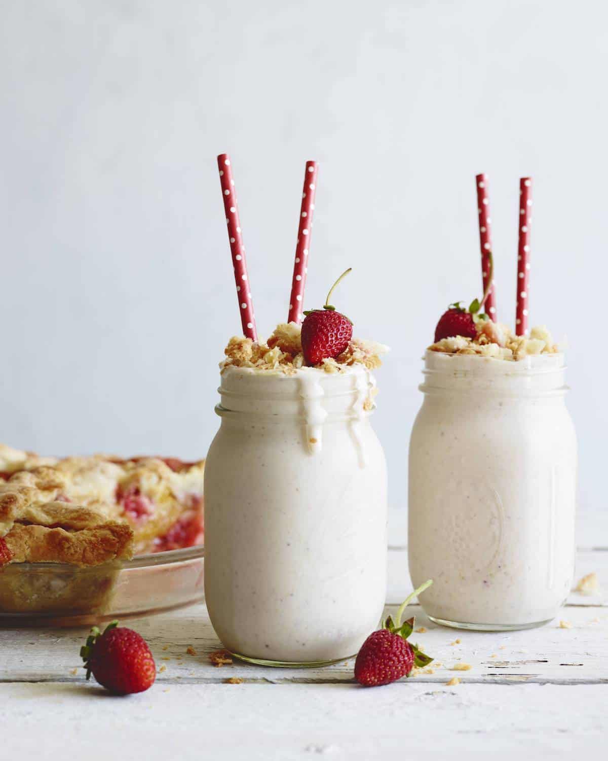 Two mason jars with strawberry milk shakes with red straws, topped with fresh strawberries and crumbled strawberry peach pie, along with a strawberry peach pie in the background.