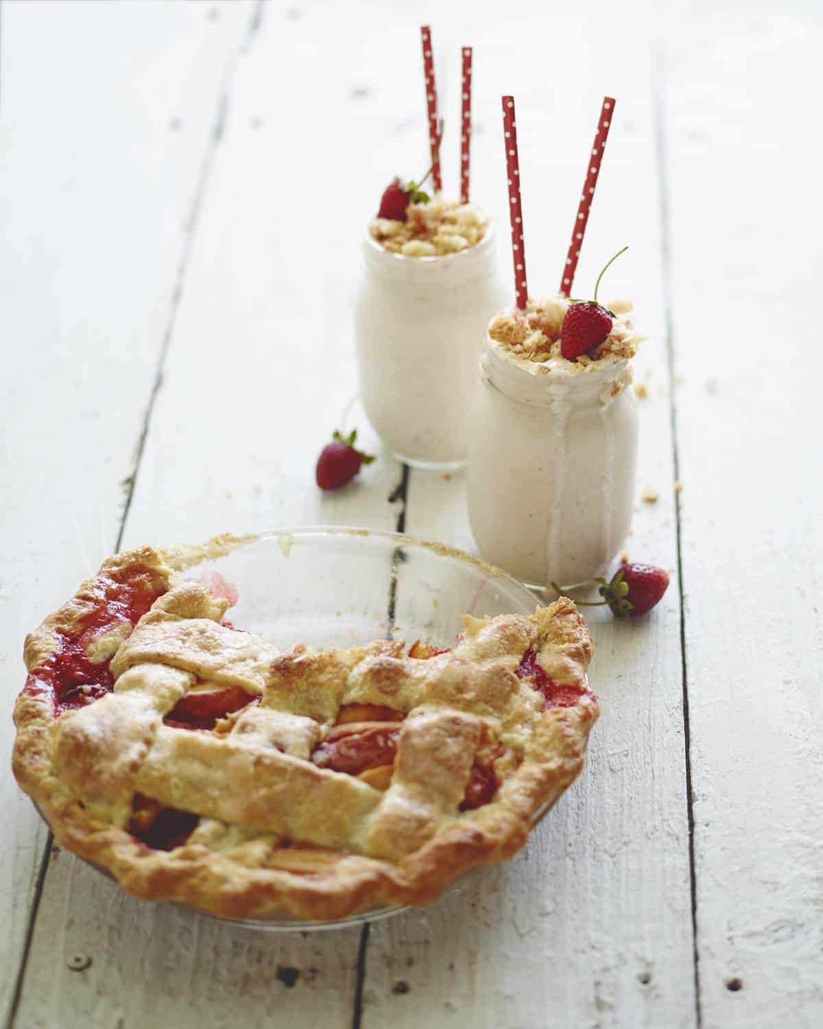 A strawberry peach pie in a glass pie dish, with a piece taken out and two mason jars with strawberry milk shakes with red straws, topped with fresh strawberries and crumbled strawberry peach pie.