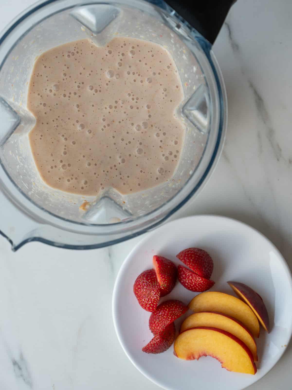 A blender with strawberry peach pie milkshake, along with a plate with peach slices and halved strawberries on the side.