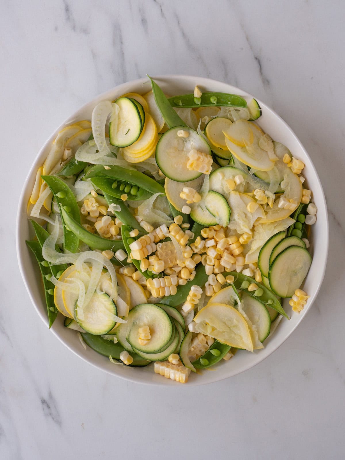 A bowl with all the veggies for raw summer salad, sliced zucchini, sliced yellow squash, corn, fennel and snap peas.