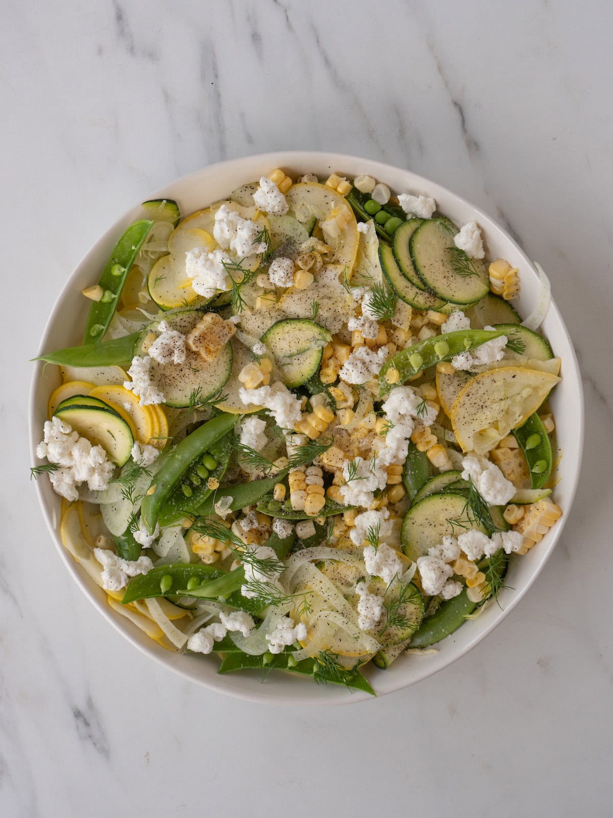 A bowl with all the veggies for raw summer salad, toped with dill, crumbled goat cheese, salt and pepper.