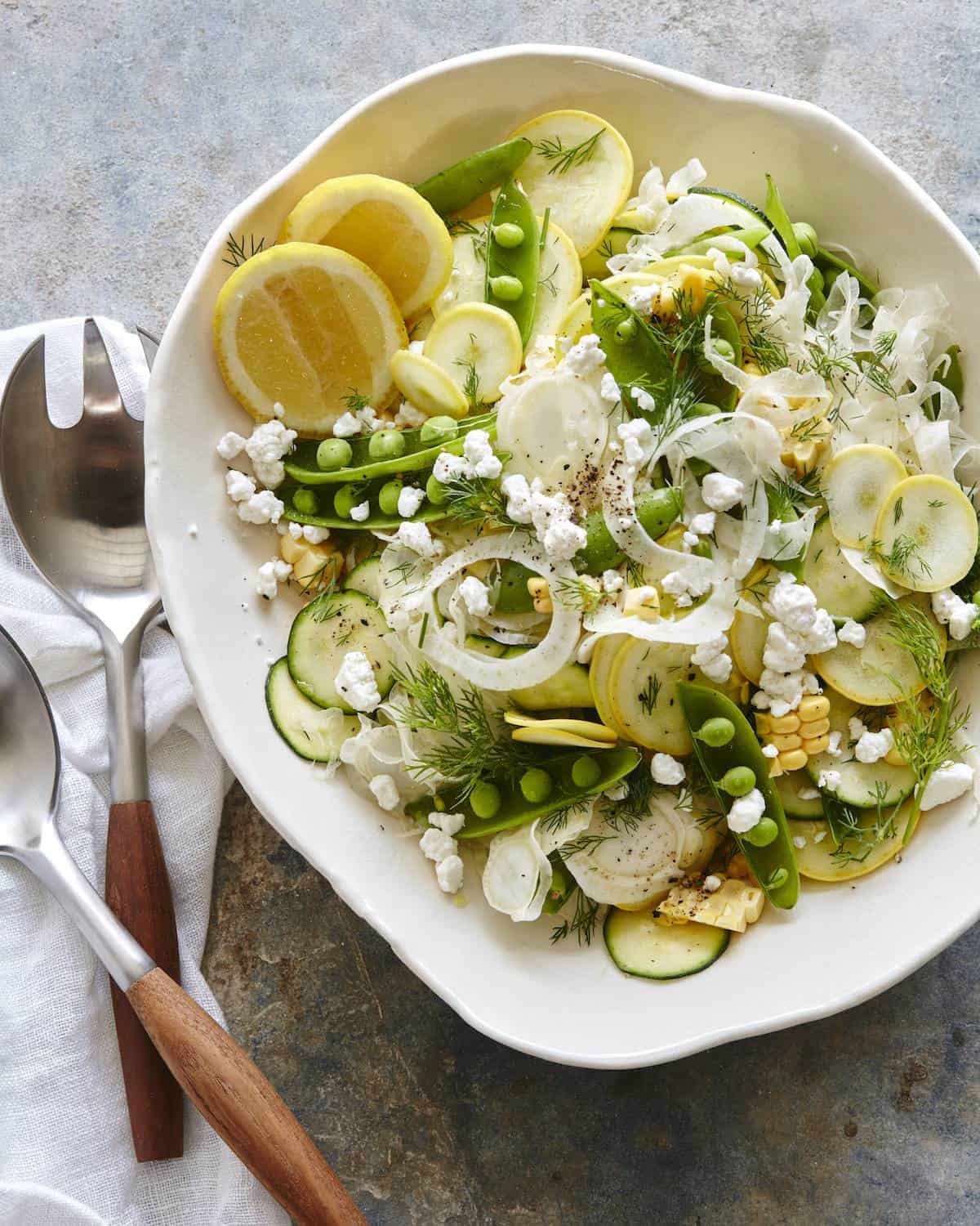 A bowl with a raw summer salad made of squash, zucchini, snap peas and fennel with lemon wedges and topped with crumbled goat cheese, along with salad spoons on the side.