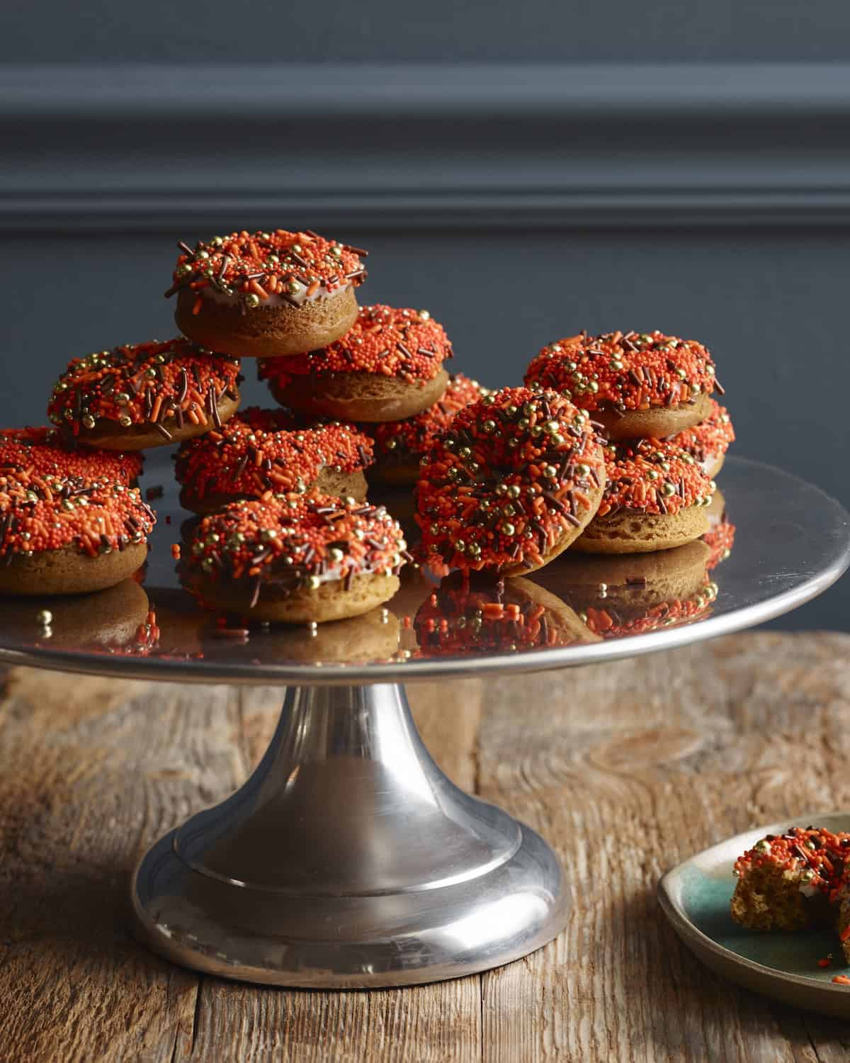 A cake stand with spiced pumpkin donuts stacked on it, and the donuts have orange, gold and chocolate sprinkles.