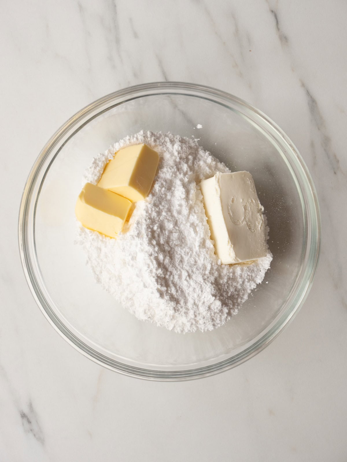 A glass mixing bowl with powdered sugar, butter and cream cheese to make icing.