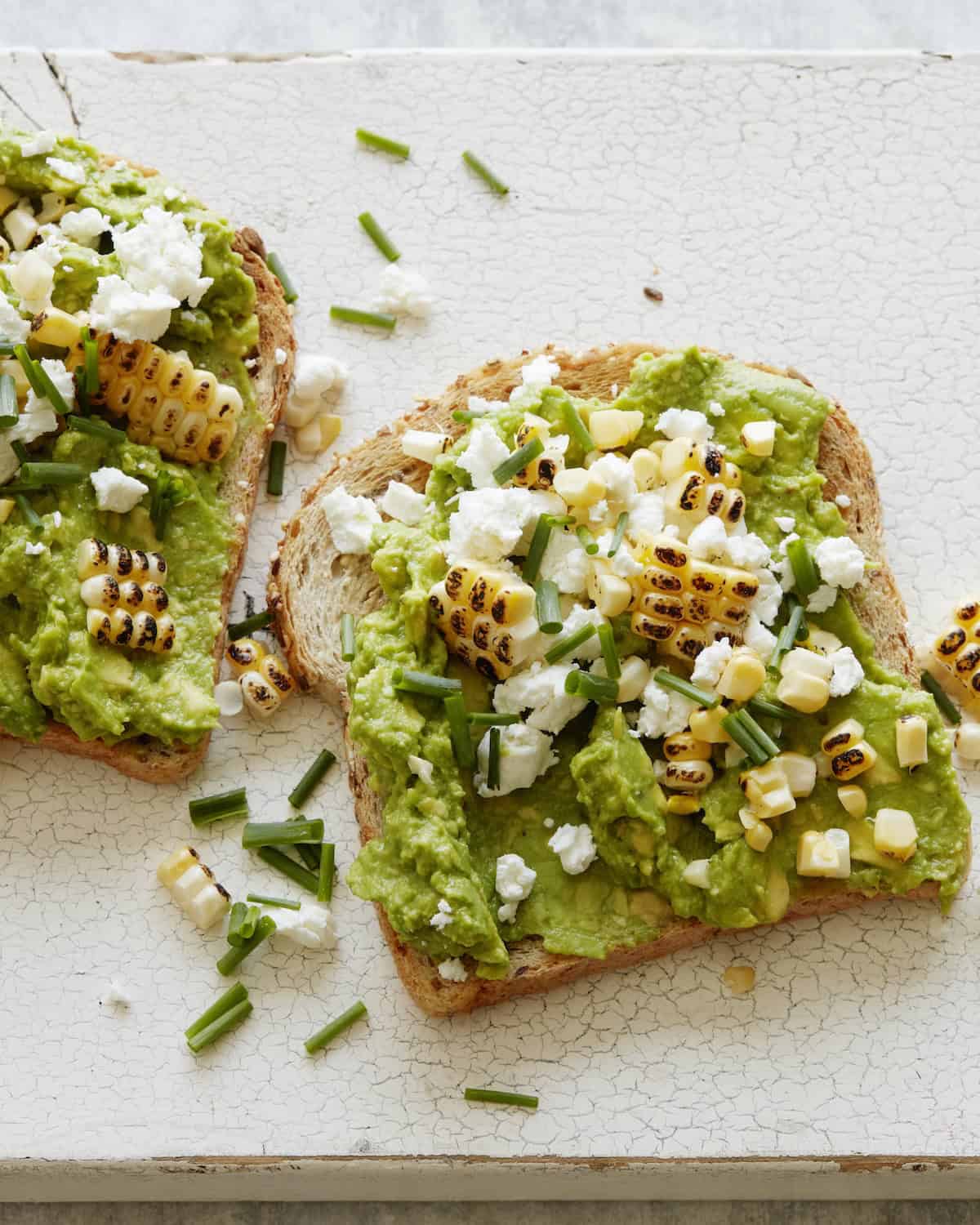 An overhead shot of two avocado toasts with crumbled goat cheese and charred corn on it, garnished with chives.