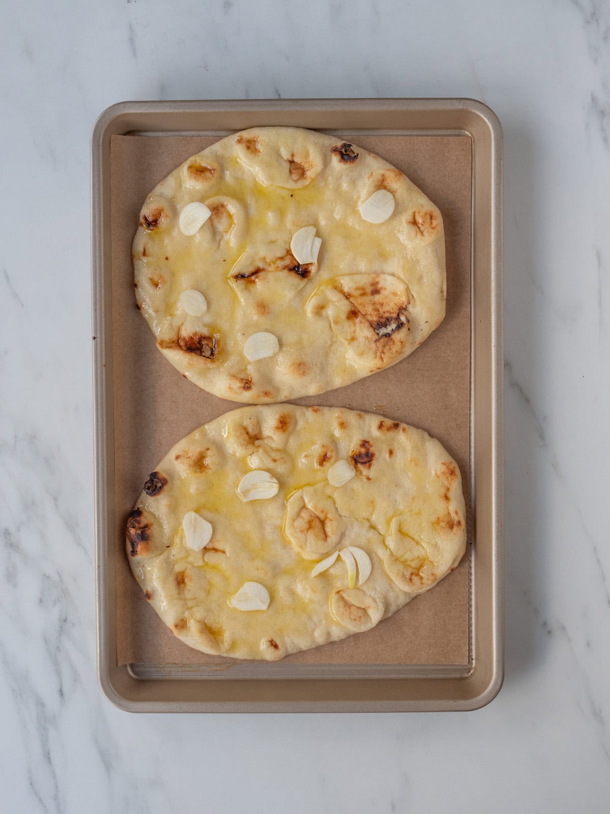 A baking sheet lined with parchment paper with two naan breads on it that are brushed with olive oil and sprinkled with slices of garlic.