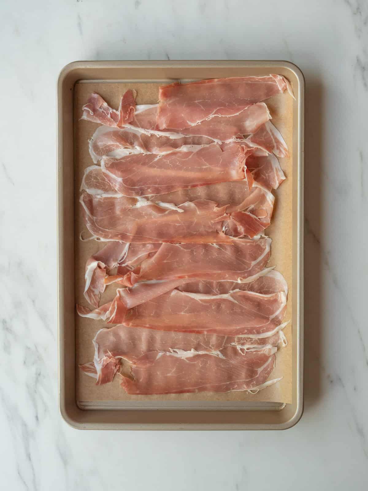 A baking sheet lined with parchment paper with prosciutto pieces placed on it, ready to be baked to crisp them up.