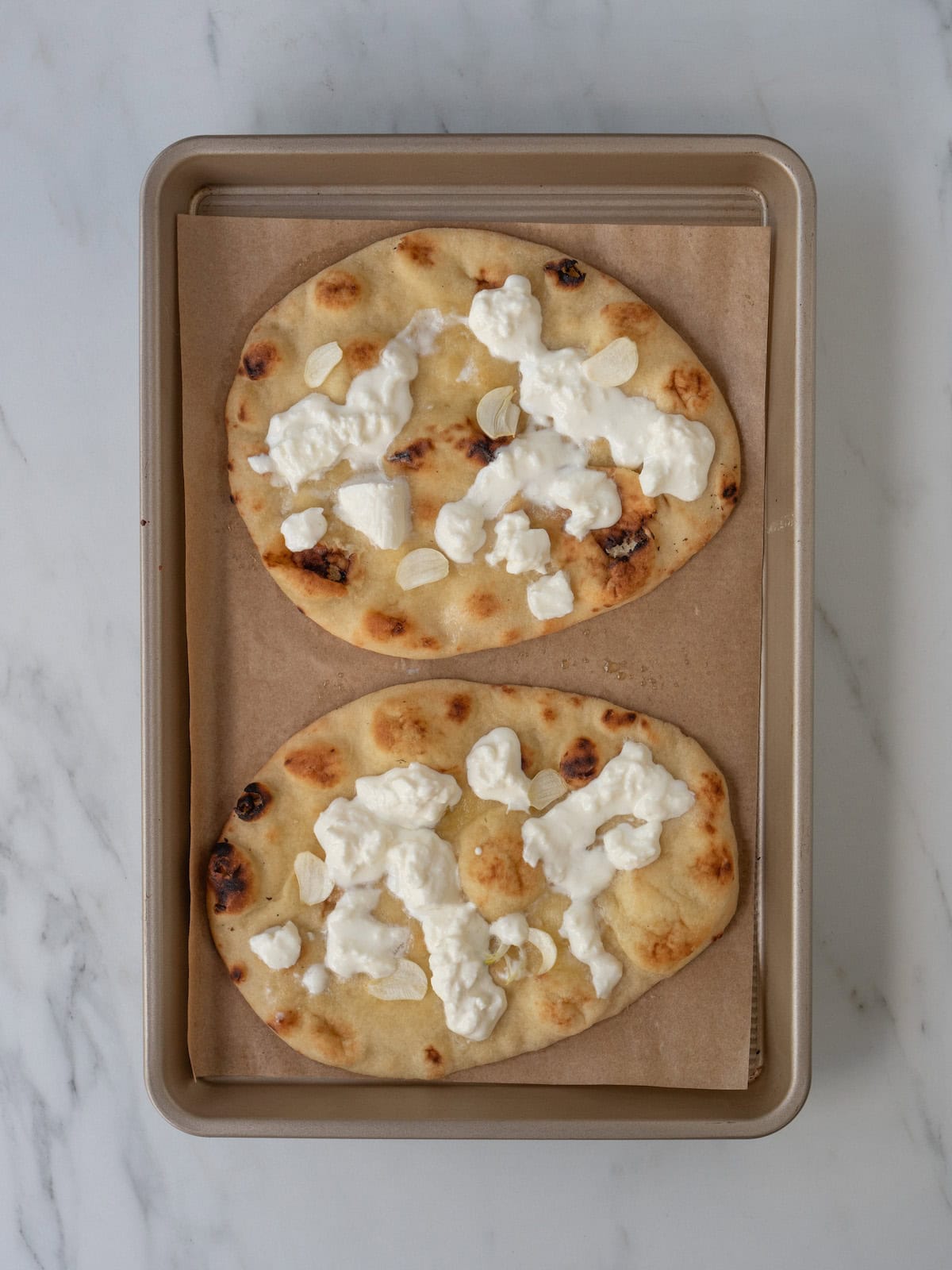 A baking sheet lined with parchment paper with two naan breads toasted in the oven and topped with burrata cheese.