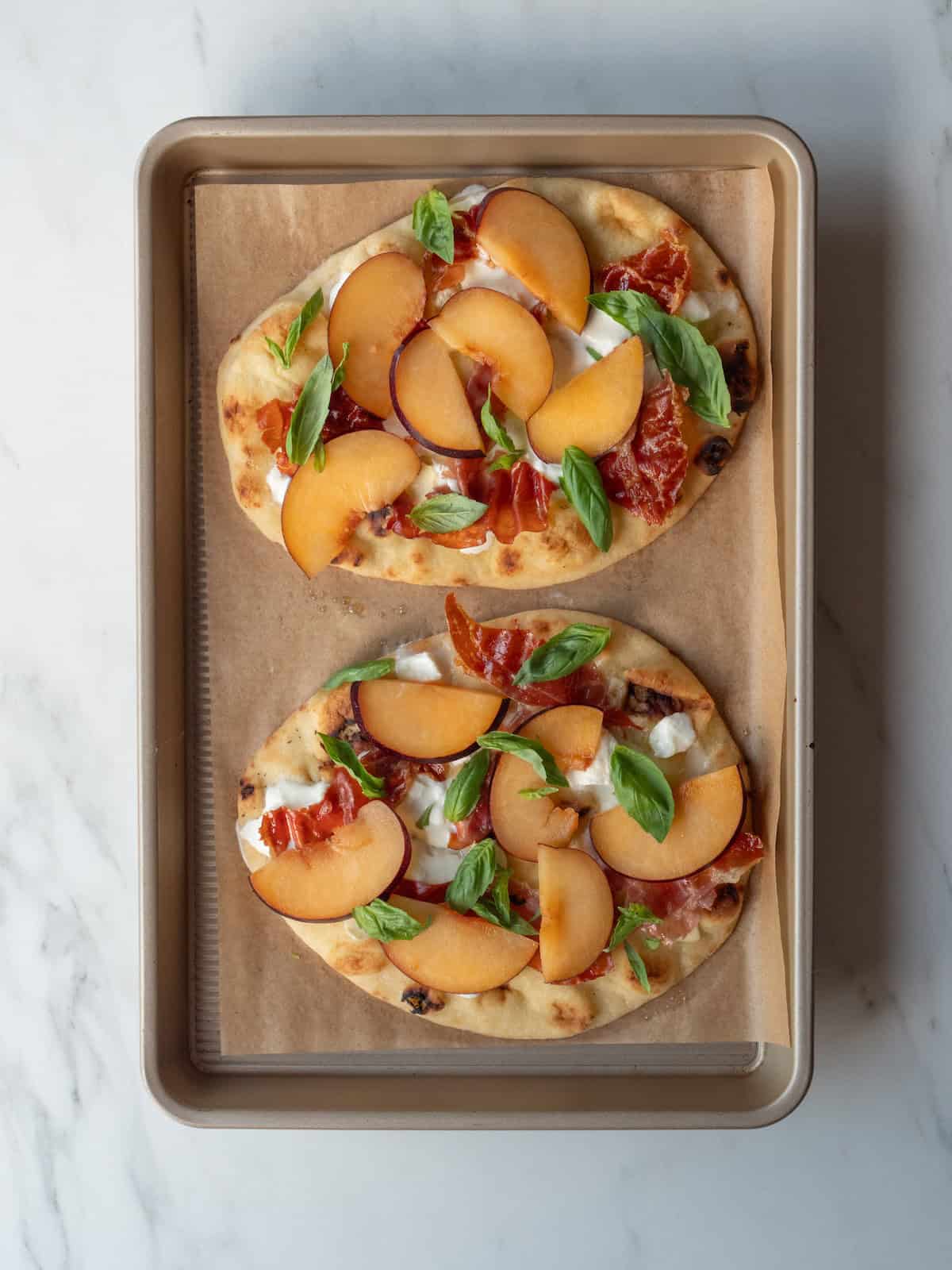 A baking sheet lined with parchment paper with two crispy prosciutto, plum and burrata flatbreads made on naan and topped with fresh basil.