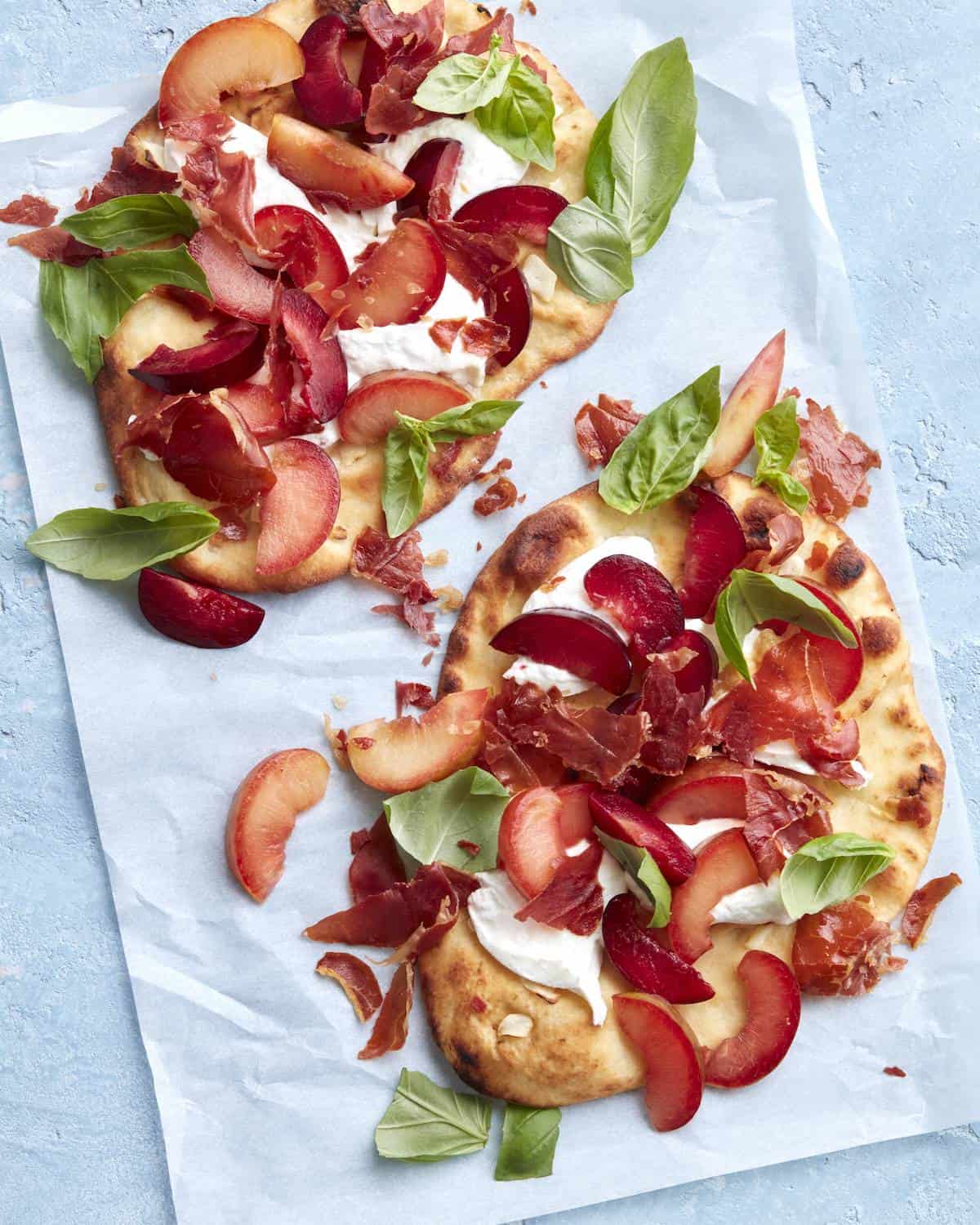 Two crispy prosciutto, plum and burrata flatbreads made on naan bread topped with fresh basil on parchment paper.