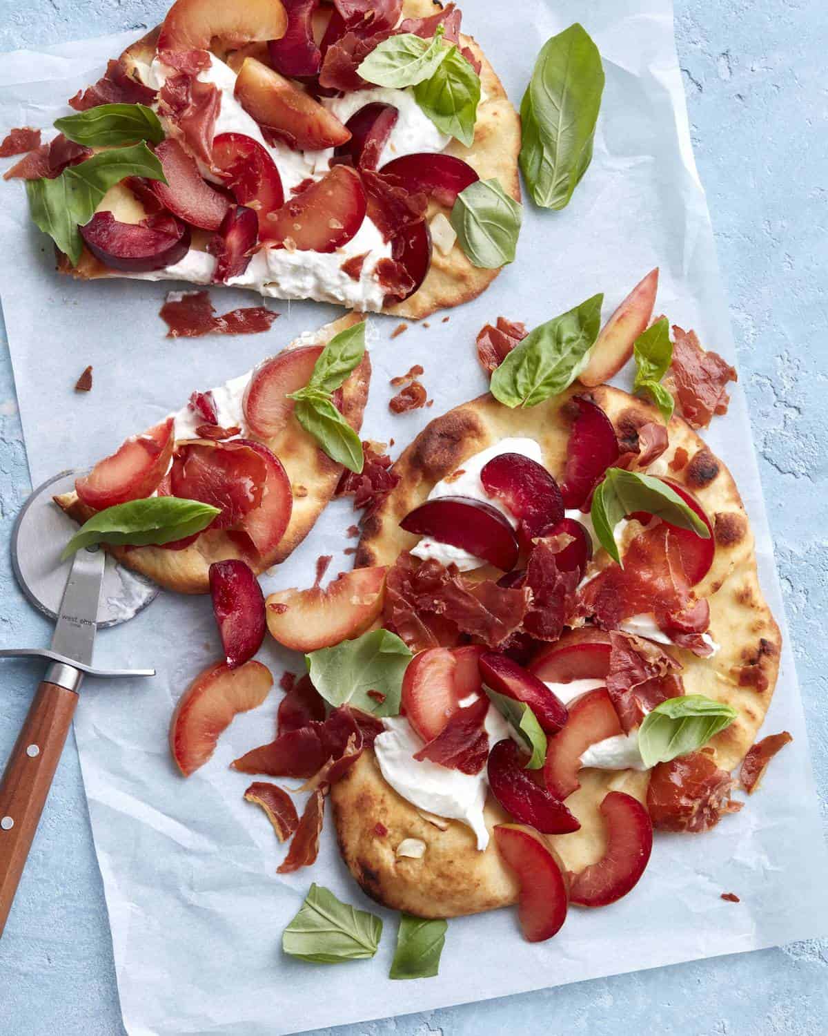 Two crispy prosciutto, plum and burrata flatbreads made on naan bread topped with fresh basil on parchment paper along with a pizza cutter.