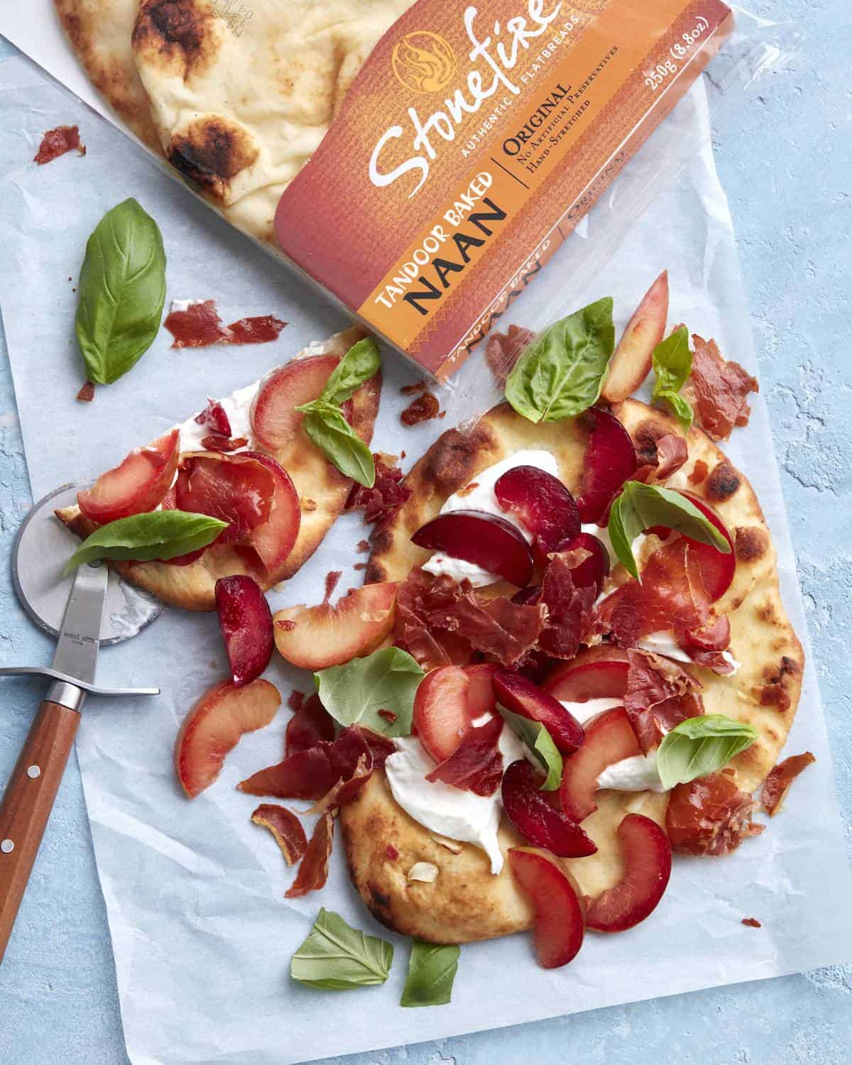 One crispy prosciutto, plum and burrata flatbread topped with fresh basil on parchment paper along with a pizza cutter and a pack of stonefire naan.