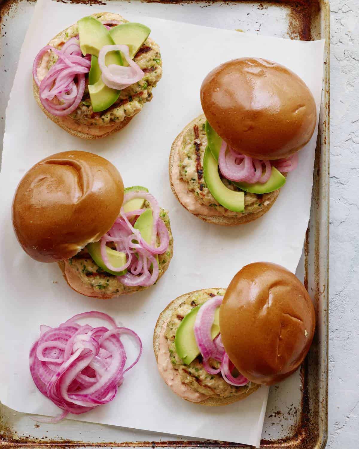 A baking sheet with parchment paper and four spiced salmon burgers with pickled red onions, harissa mayo and avocado.