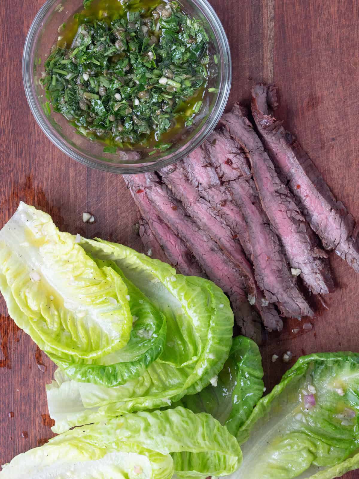 An overhead shot of a wooden board with sliced grilled flank steak cooked medium along with a bowl of salsa verde and baby romaine leaves.