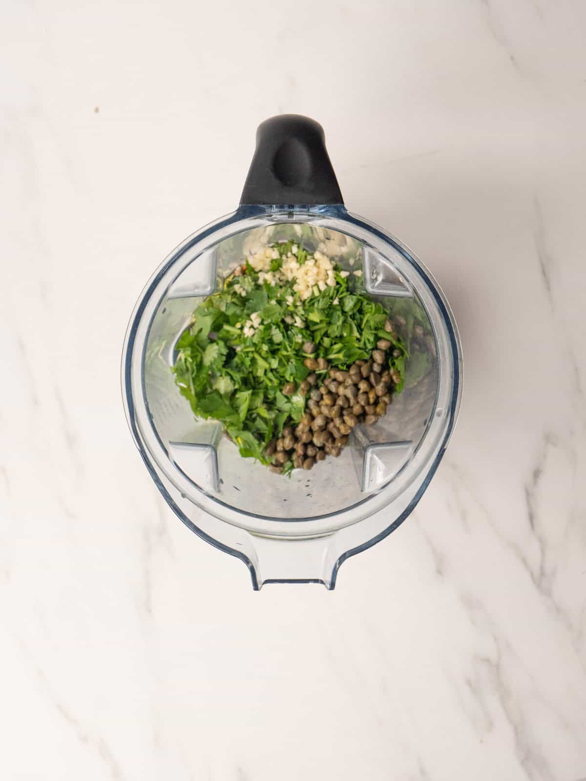 A blender jar with herbs, garlic, vinegar and capers to make salsa verde.