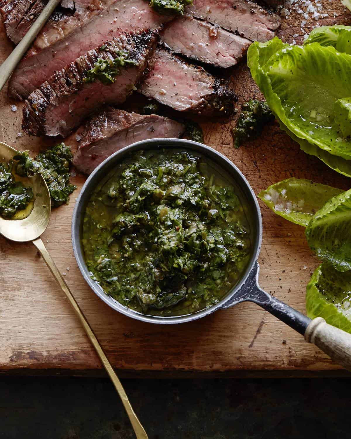 A closeup shot of a wooden board with sliced grilled flank steak cooked medium with salsa verde drizzled on it, along with a bowl of salsa verde and baby romaine leaves.