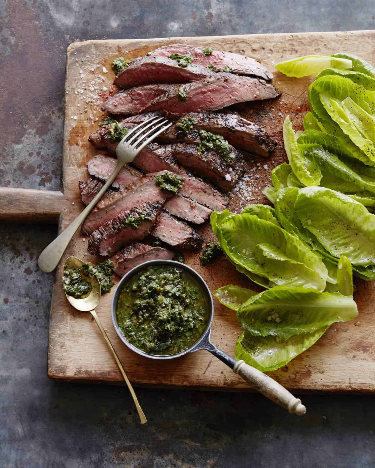 A wooden board with sliced grilled flank steak cooked medium with salsa verde drizzled on it, along with a bowl of salsa verde and baby romaine leaves.