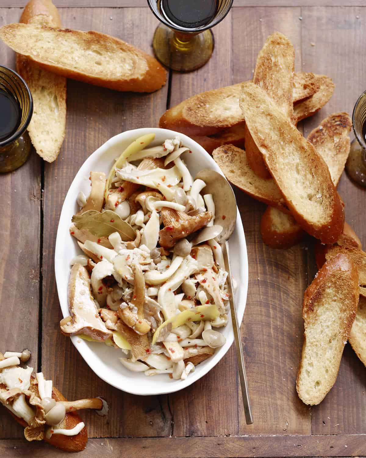 A low bowl with marinated mushrooms along with toasted bread on the side along with two beverages.