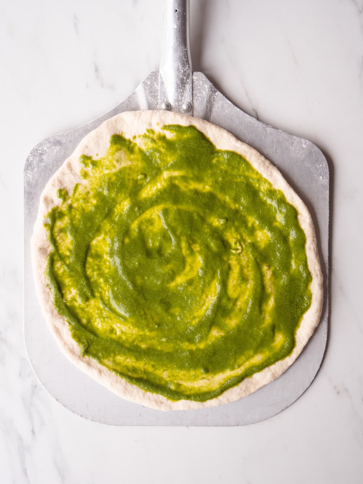 A pizza peel with a stretched out pizza dough spread with basil vinaigrette.