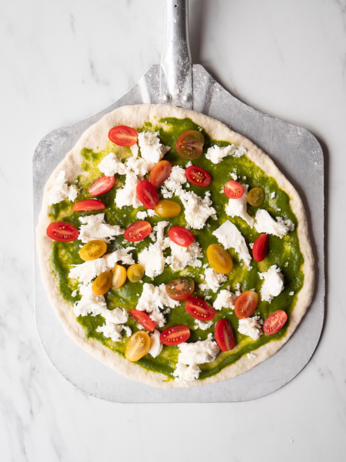 A pizza peel with a stretched out pizza dough spread with basil vinaigrette and topped with halved cherry tomatoes and torn pieces of mozzarella cheese.
