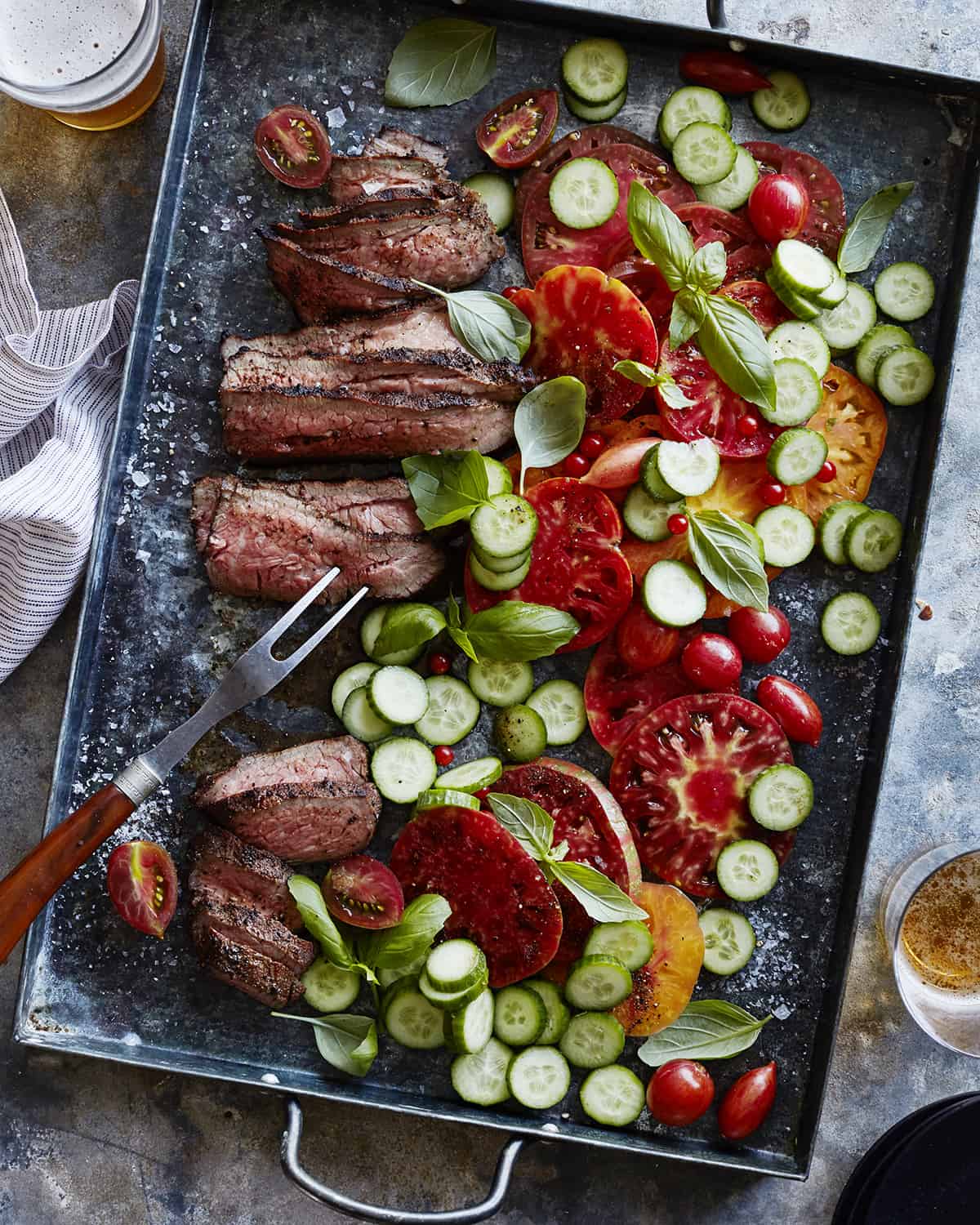 Cooked Tri Tip on a sheet pan with sliced tomatoes, sliced cucumbers and a butchers fork.