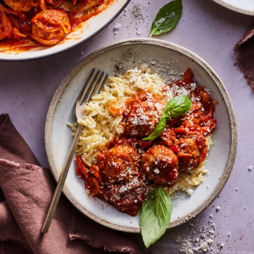 A braiser with chicken meatballs in red sauce with a garnish of basil on a purple table. On the table there is also a small ceramic bowl of creamy orzo and another small ceramic bowl with a serving of chicken meatballs over creamy orzo with a gold fork and two mauve linen napkins.