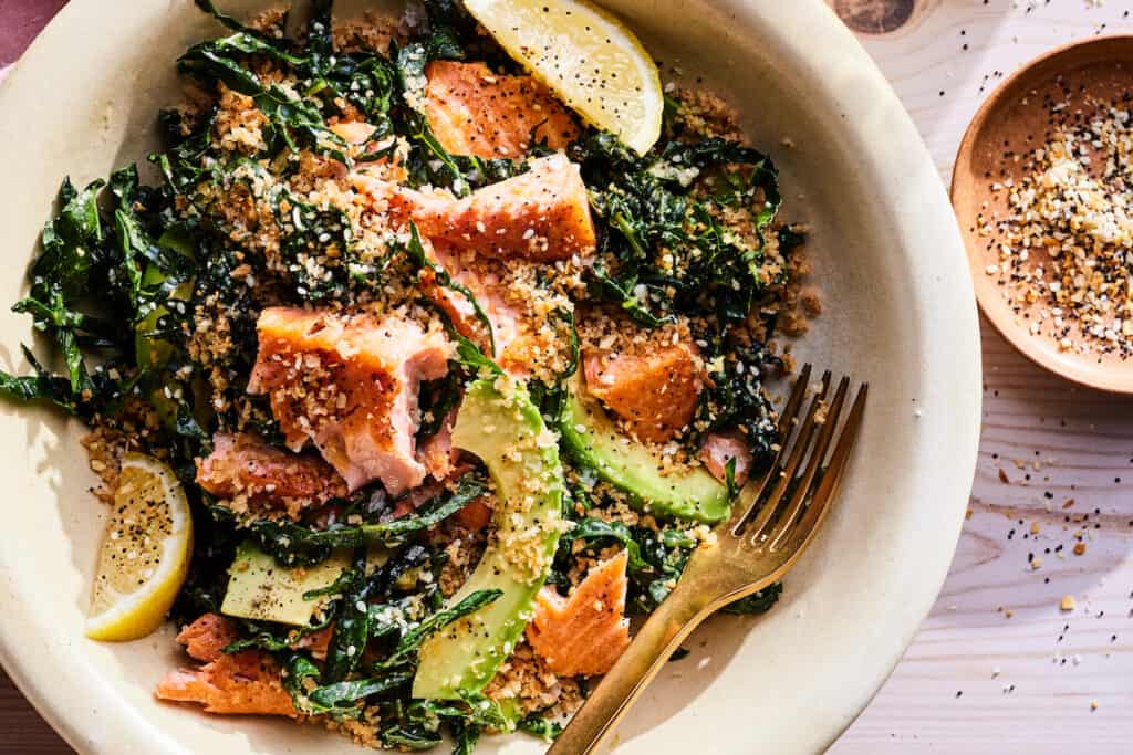 An overhead shot of a beige ceramic bowl with salmon kale salad on a light wooden table. The kale salad has flaked pieces of crispy salmon sliced of avocados and lemons and a healthy garnish of everything but the bagel seasoning. A gold fork is resting in the bottom right edge of the bowl.