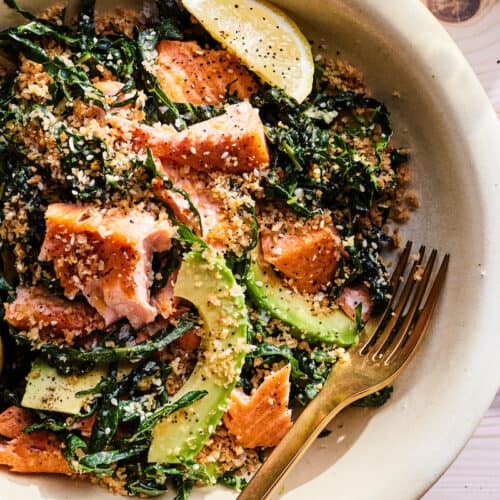An overhead shot of a beige ceramic bowl with salmon kale salad on a light wooden table. The kale salad has flaked pieces of crispy salmon sliced of avocados and lemons and a healthy garnish of everything but the bagel seasoning. A gold fork is resting in the bottom right edge of the bowl.