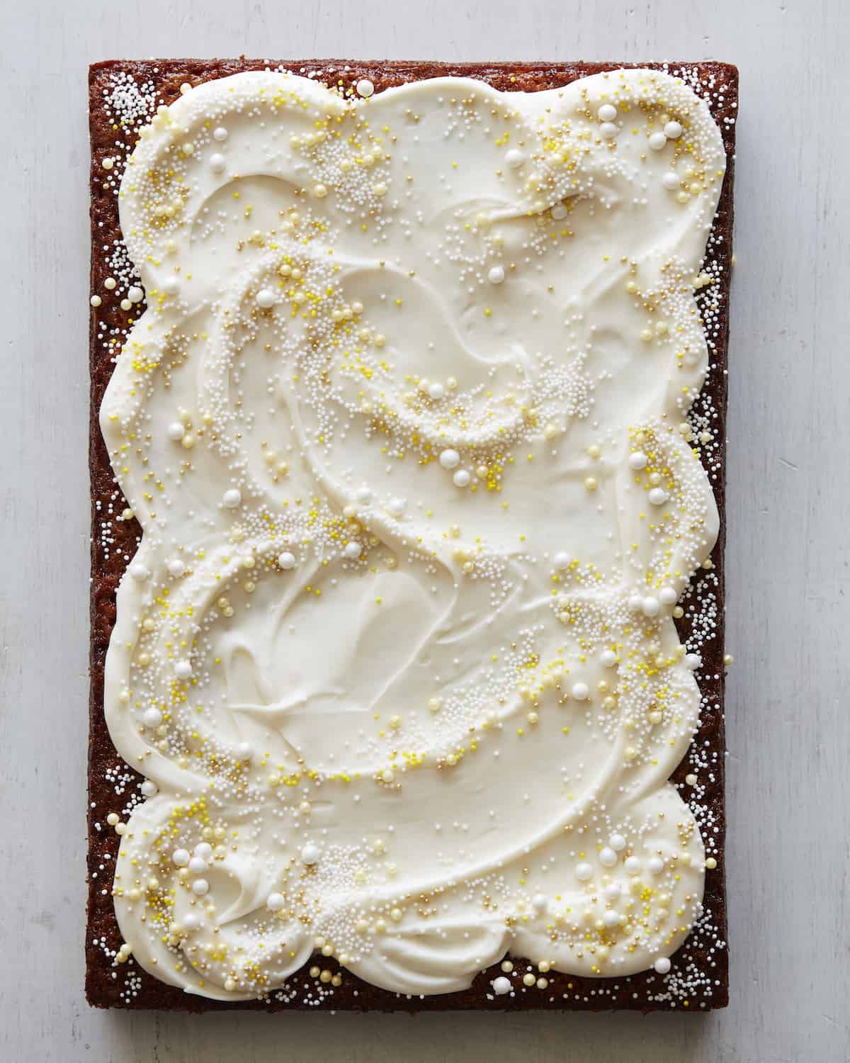 A rectangle Pineapple Sheet Cake with big fluffy swirls of white cream cheese frosting and yellow and white nonpareils and pearl sprinkles on a grey background