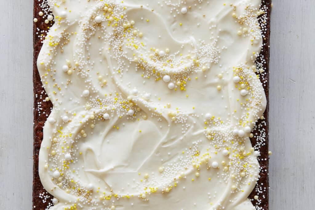 A rectangle Pineapple Sheet Cake with big fluffy swirls of white cream cheese frosting and yellow and white nonpareils and pearl sprinkles on a grey background