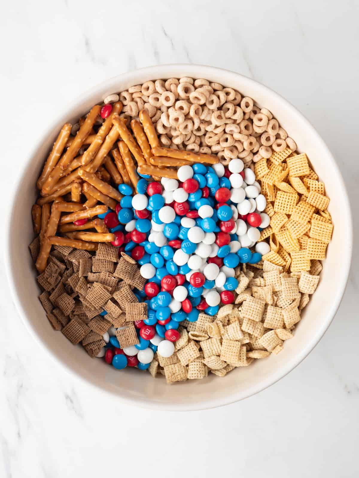 A bowl with cheerios, corn chex, wheat chex, rice chex, pretzels and M&Ms. 