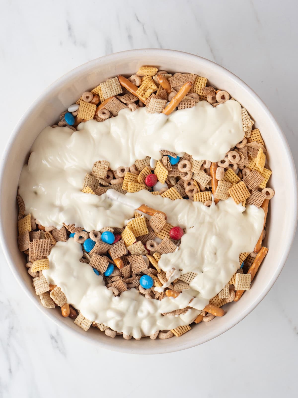 A bowl with a mix of chex, M&Ms, pretzels and cheerios with molten white chocolate poured on it.
