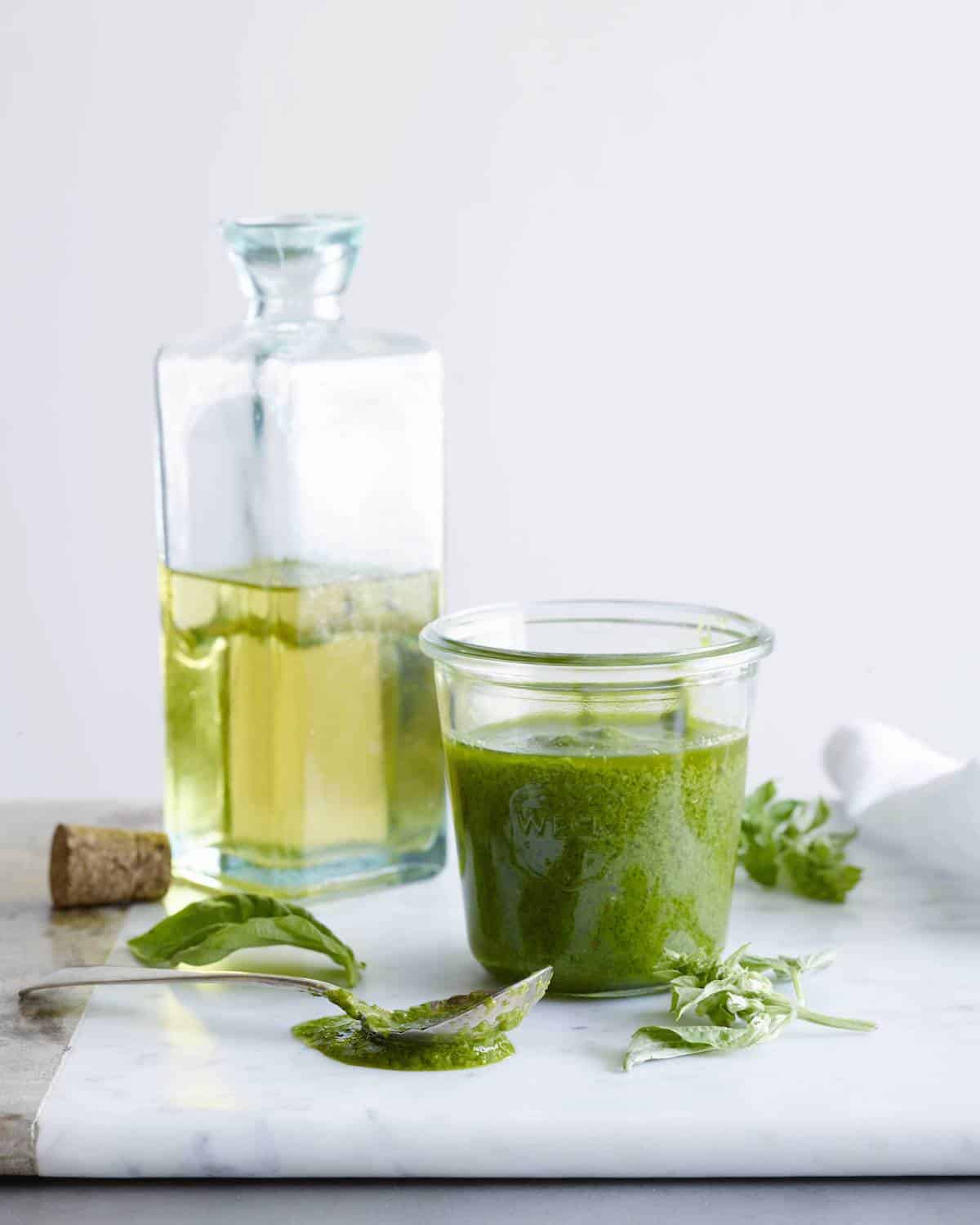 basil vinaigrette recipe in a weck jar with a square glass olive oil bottle in the background, a few basil leaves on the table with a spoon dipped in basil vinaigrette on the table