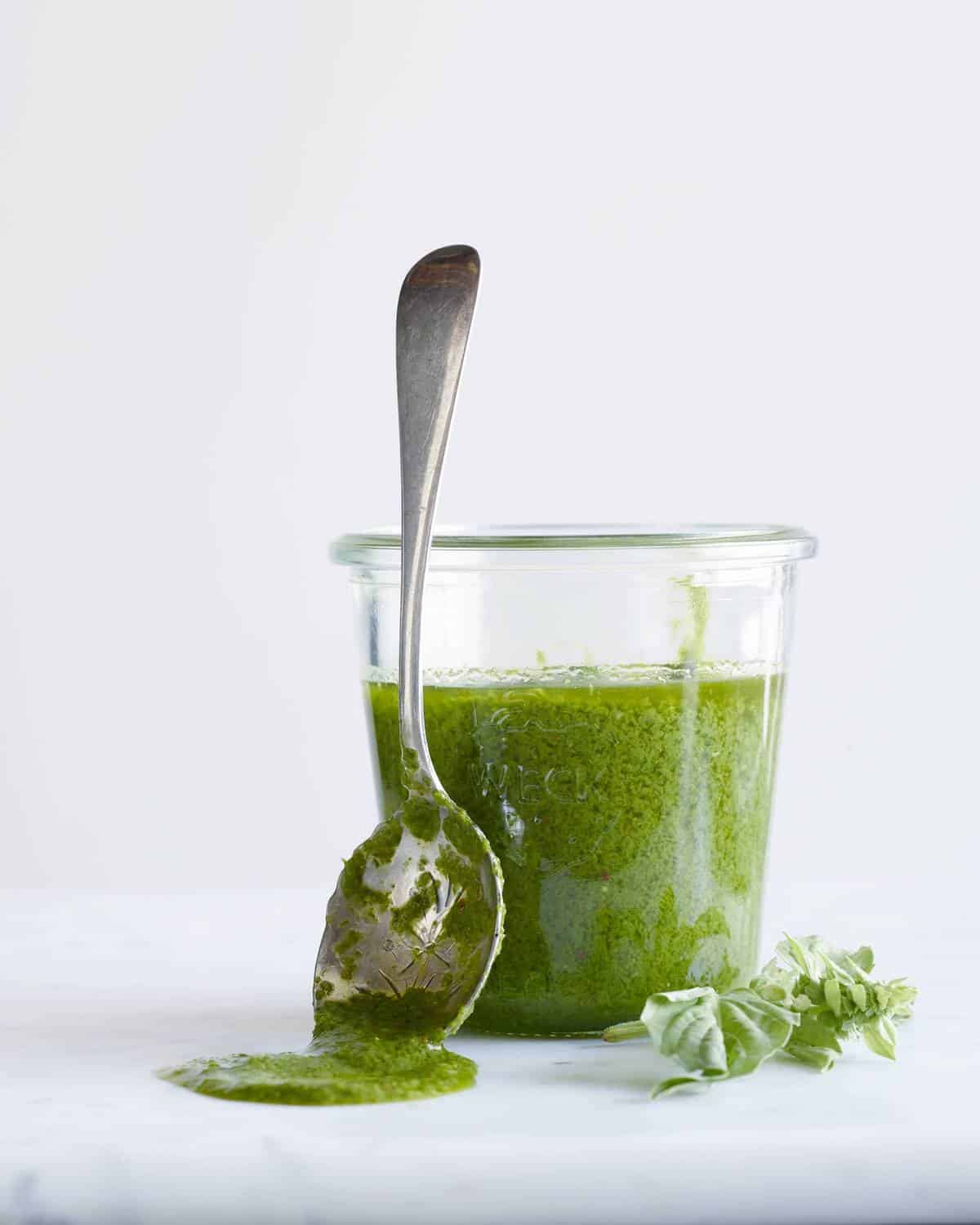 Basil vinaigrette in a weck jar with a spoon covered in vinaigrette propped up on the jar and a sprig of basil in bottom right corner.
