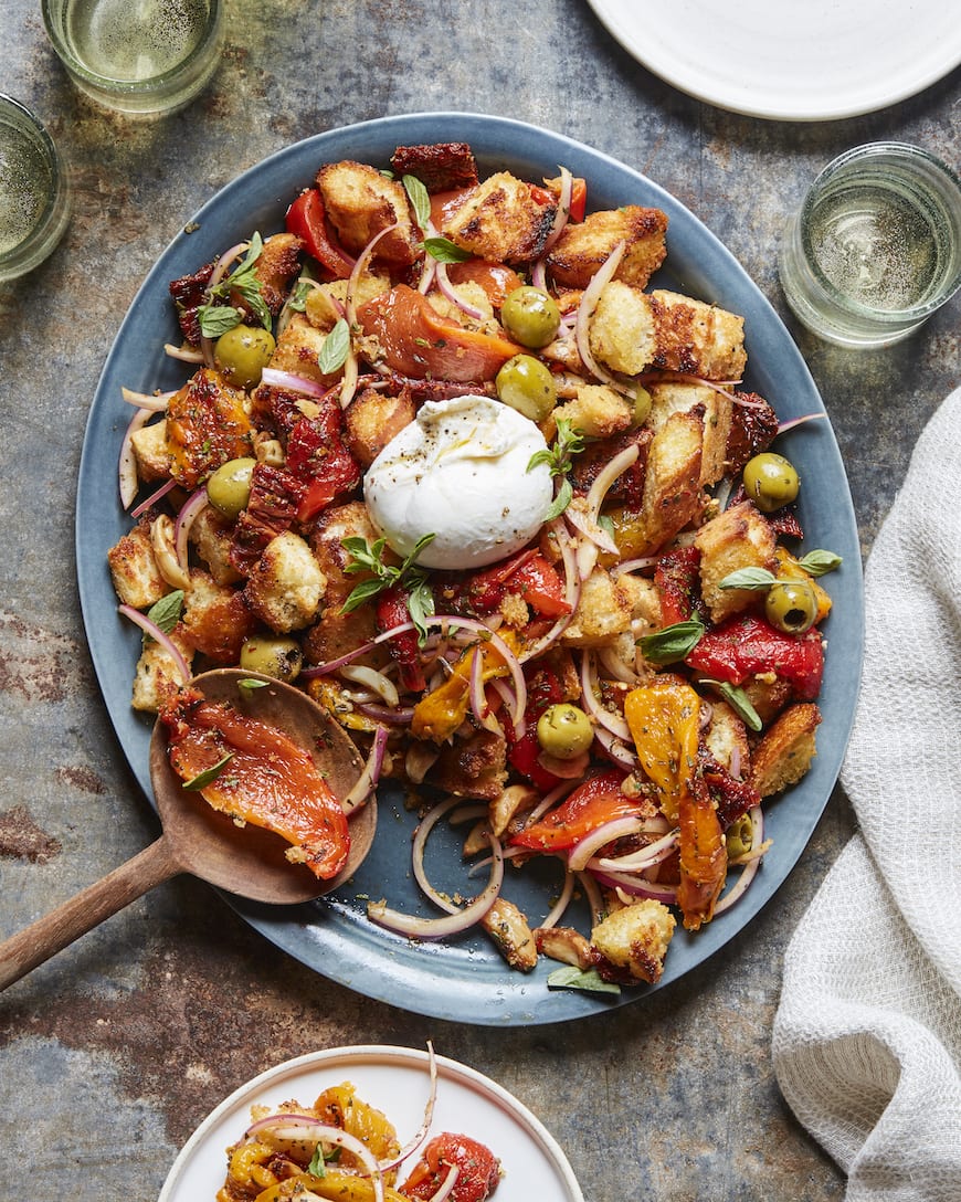 A blue oval platter with burrata panzanella, with roast peppers olives and onions, a wooden spoon to serve and a plate with some panzanella as well as some glasses with a drink on the side.
