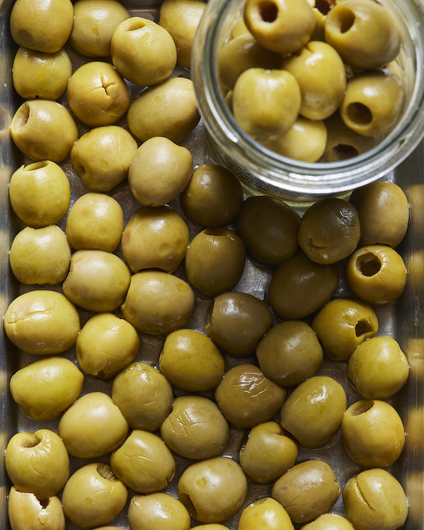 Closeup shot of a glass jar of green olives placed in a flat platter full of green olives.