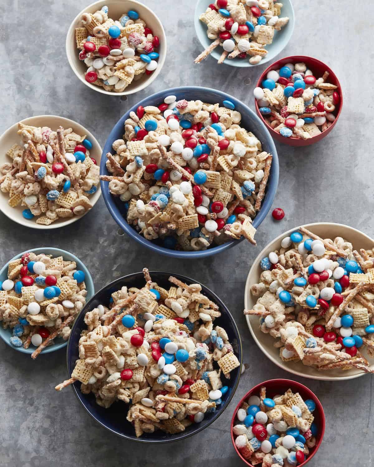 An overhead shot of bowls of different sizes with 4th of July mix made up of a variety of cheerios, chex, pretzels and M&Ms mixed in white chocolate.