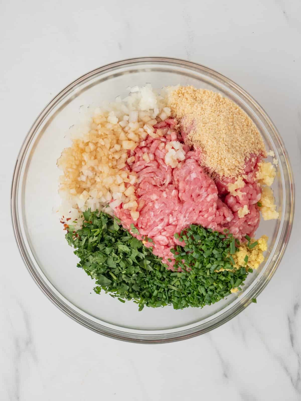 A large glass mixing bowl with ground pork, seasonings and finely chopped ginger, garlic and onion, seasonings and bread crumbs to make asian pork meatballs.