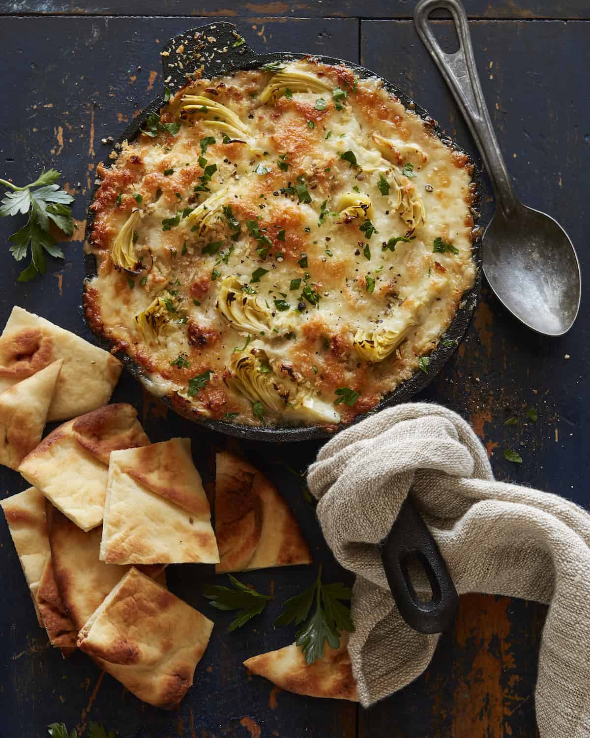 A skillet with cheesy artichoke dip and some cut up naan bread on its side, a spoon and a dish cloth wrapping up the skillet's handle.
