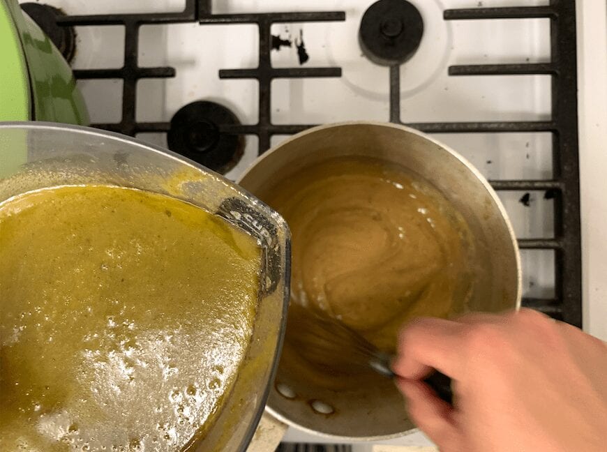 A pan with a roux made with turkey drippings, and stock being poured in while a hand whisking as the stock is poured in.