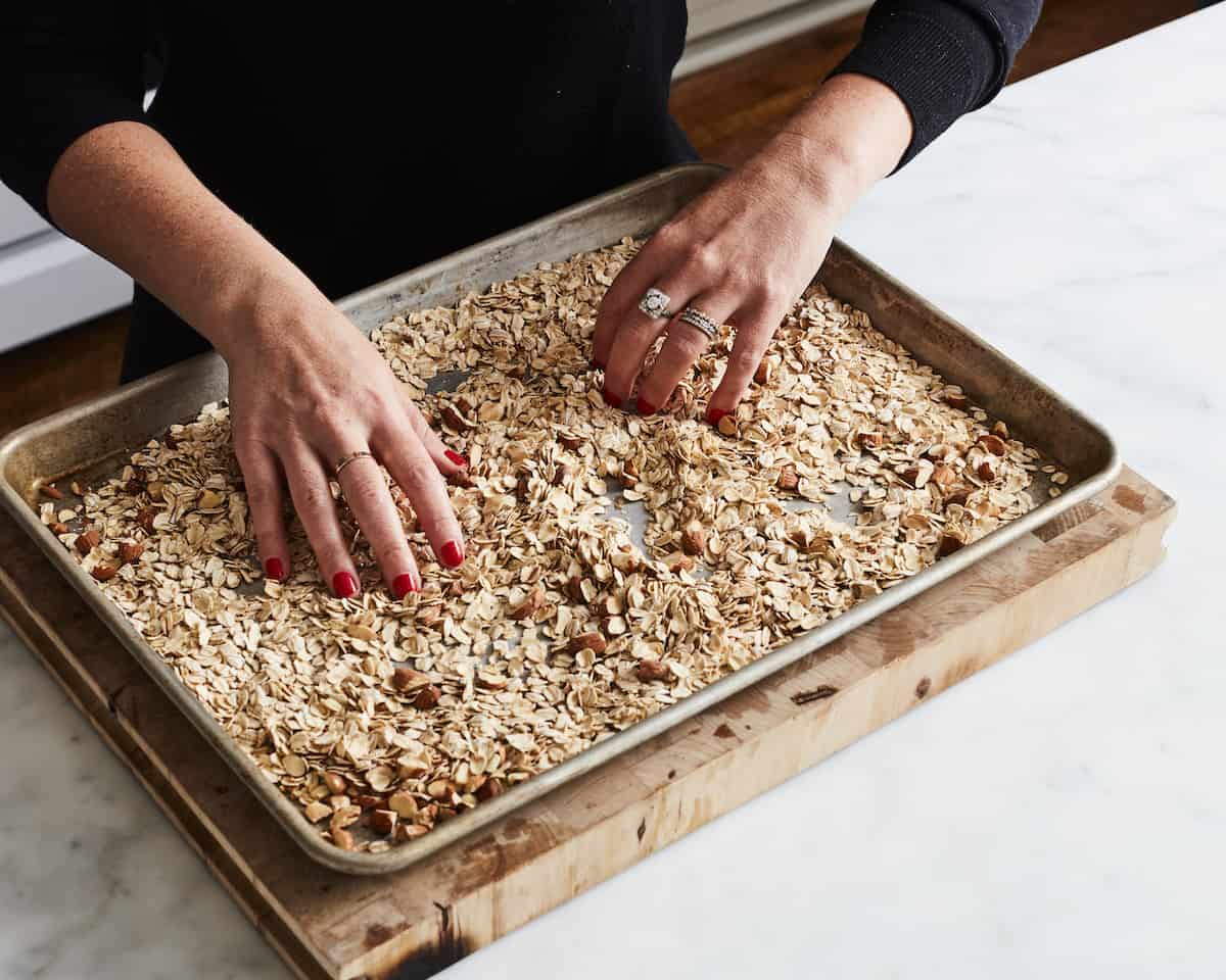 A woman's hands mixing oats and almonds on a baking sheet placed on a wooden chopping board.