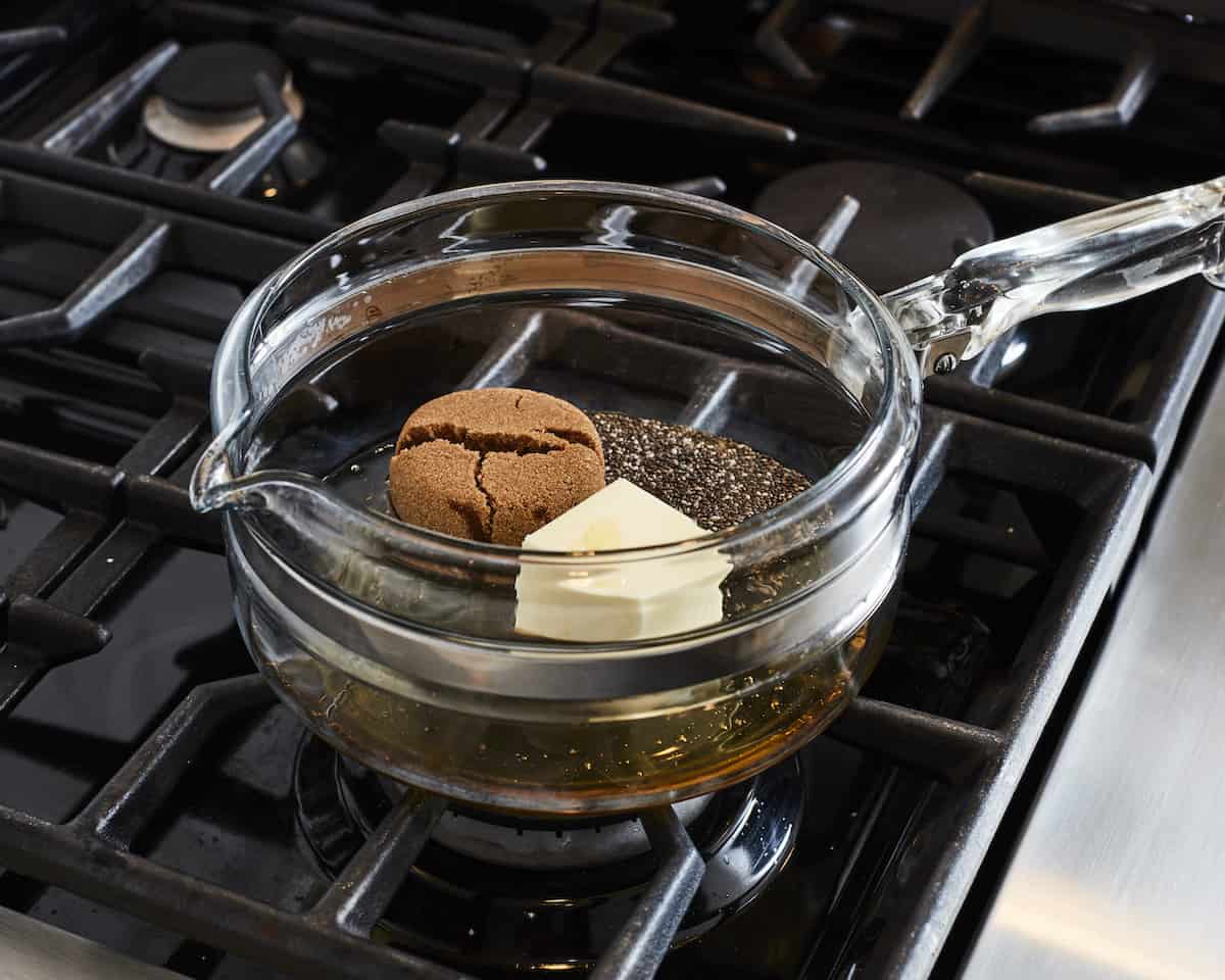 A small glass saucepan on the stove with butter, honey, brown sugar, vanilla extract, chia and salt all placed in it.