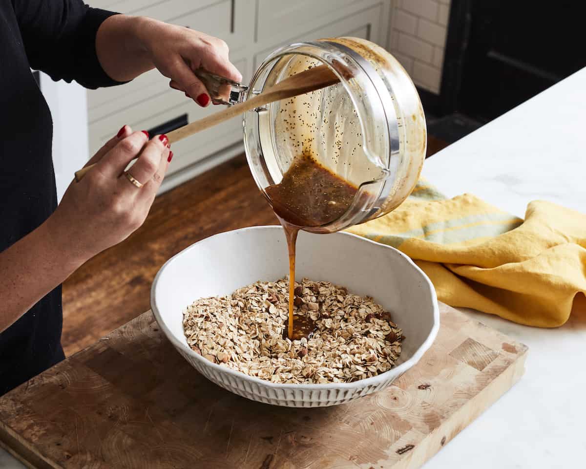 A woman pouring a smooth butter, honey and brown sugar mixture from a small saucepan into a white ceramic bowl with toasted almonds and oats placed on a wooden chopping board.