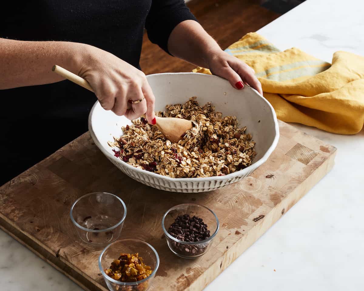 A woman mixing together cranberries and raisins into an oats, almonds and butter, honey mixture using a wooden spoon, with three small glass bowls with the cranberries, golden raisins and chocolate chips on the side.